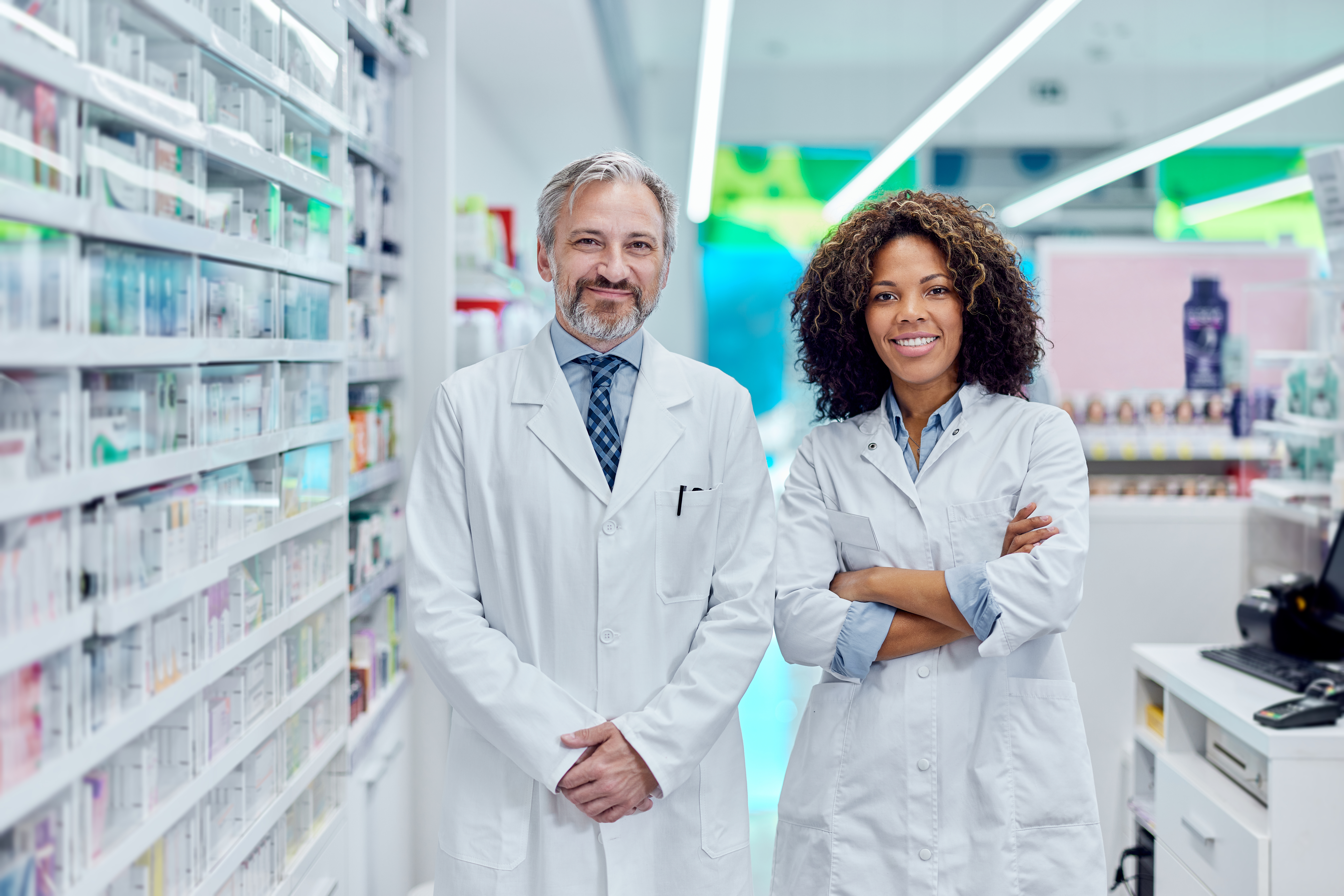 Pharmacists Standing in Modern Pharmacy ‣ DLM Connections Pharmaceutical partners connected through DLM Connections standing in a modern pharmacy.